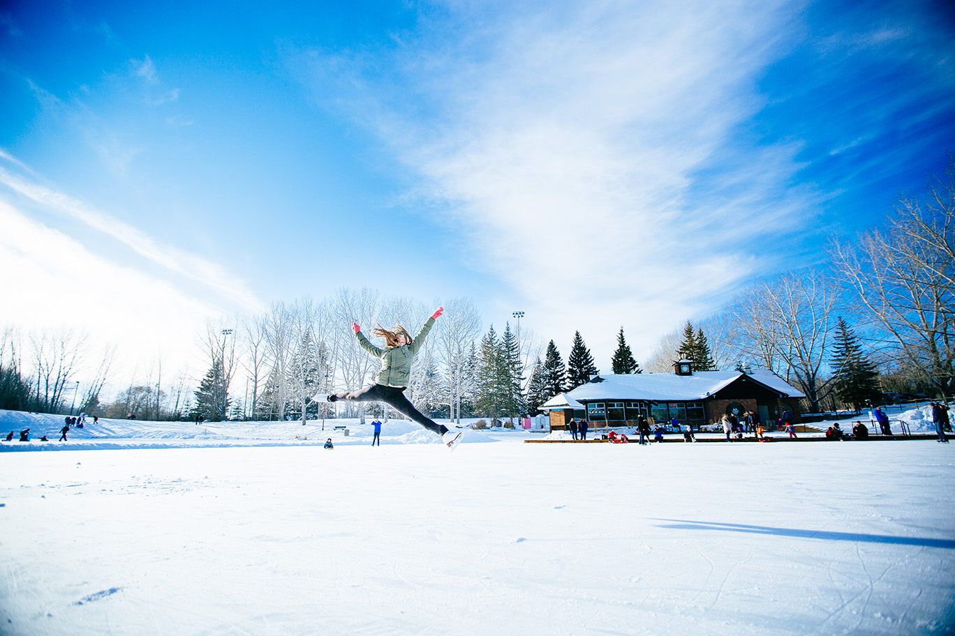 Figure skater does a split jump on the ice in front of the Bower Ponds pavilion agains a blue sky with whispy clouds