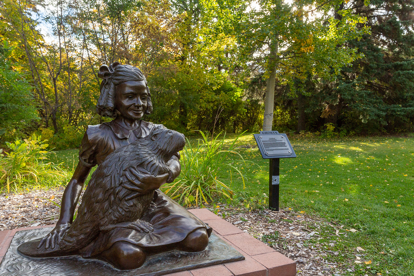 Metal Ghost statue of Micky the Beaver being held by a kneeling Doris Forbes in a green glen of trees in Cornation Park