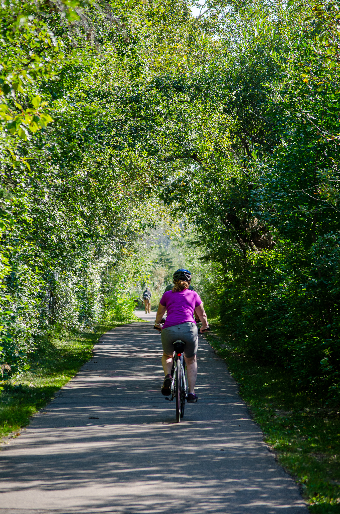 Biker in a bright blue coat riding their bike next to a smooth pond with a fountain in it reflecting the trees around it