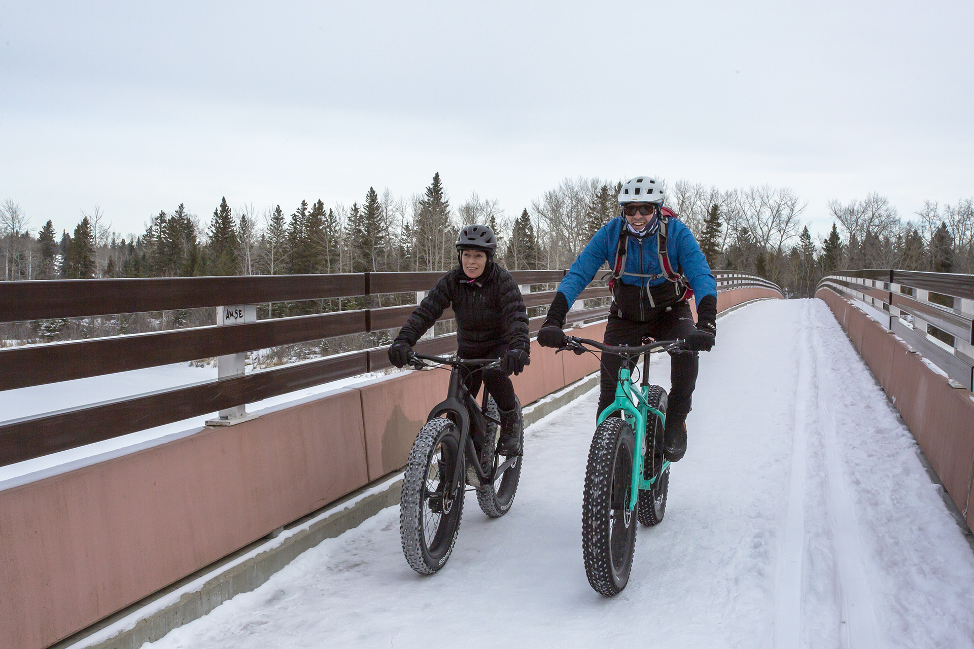 Two people fat tire biking across bridge at heritage ranch in red deer in the winter time.